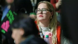 Eine Frau mit großer Brille sitzt aufmerksam in einer Menschenmenge bei einer Veranstaltung.