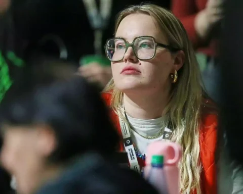 Eine Frau mit großer Brille sitzt aufmerksam in einer Menschenmenge bei einer Veranstaltung.