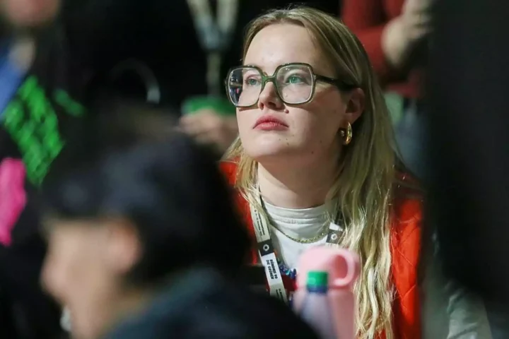 Eine Frau mit großer Brille sitzt aufmerksam in einer Menschenmenge bei einer Veranstaltung.