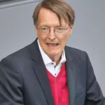 Karl Lauterbach spricht vor grauem Hintergrund und trägt einen dunklen Anzug sowie einen roten Pullover.