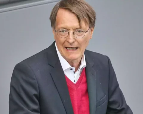 Karl Lauterbach spricht vor grauem Hintergrund und trägt einen dunklen Anzug sowie einen roten Pullover.