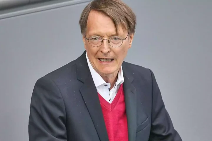 Karl Lauterbach spricht vor grauem Hintergrund und trägt einen dunklen Anzug sowie einen roten Pullover.