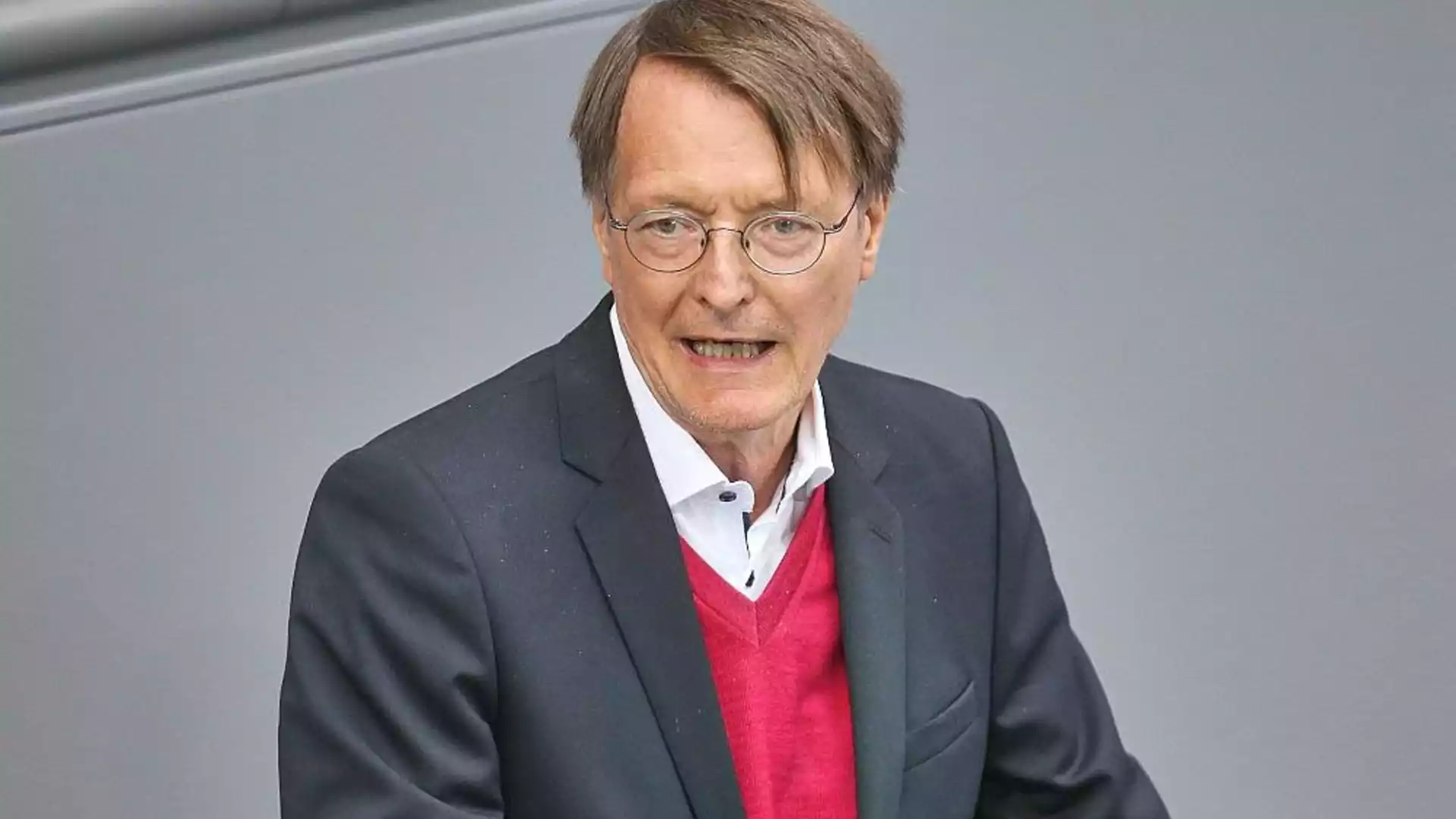 Karl Lauterbach spricht vor grauem Hintergrund und trägt einen dunklen Anzug sowie einen roten Pullover.