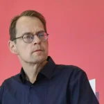 Ein Mann mit Brille steht vor einem Mikrofon vor rosafarbenem Hintergrund.