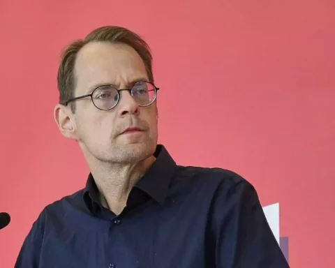 Ein Mann mit Brille steht vor einem Mikrofon vor rosafarbenem Hintergrund.