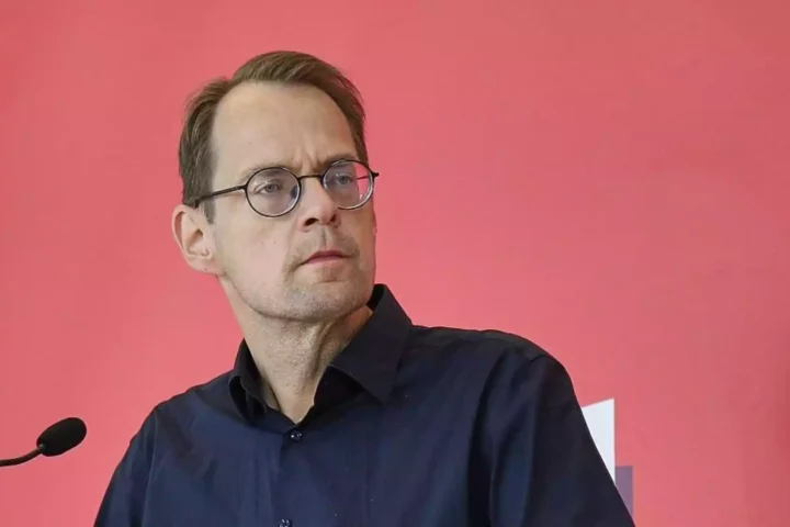 Ein Mann mit Brille steht vor einem Mikrofon vor rosafarbenem Hintergrund.