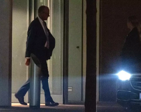 Mann im Anzug geht nachts an einem beleuchteten Gebäude vorbei, rechts steht ein Wagen mit eingeschaltetem Scheinwerfer.