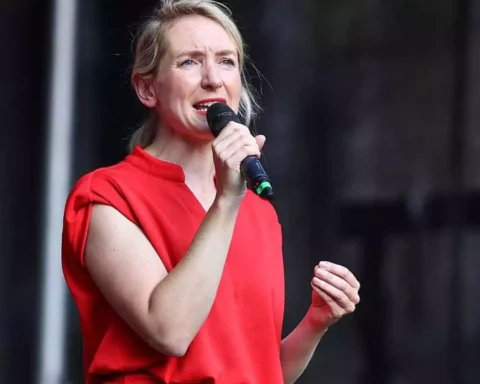Ines Schwerdtner spricht mit einem Mikrofon auf einer Bühne und trägt ein rotes Oberteil.
