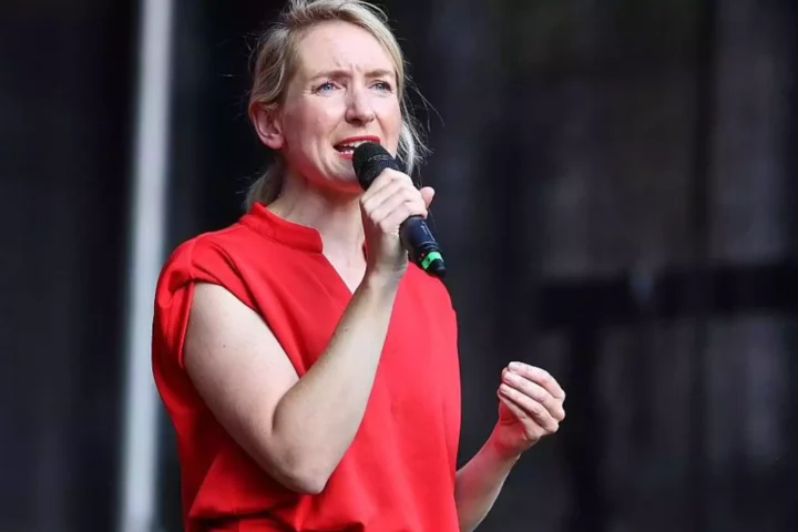 Ines Schwerdtner spricht mit einem Mikrofon auf einer Bühne und trägt ein rotes Oberteil.