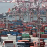 Containerhafen mit gestapelten Containern, Hafenkränen und Frachtschiffen im Hintergrund.