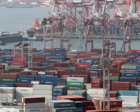 Containerhafen mit gestapelten Containern, Hafenkränen und Frachtschiffen im Hintergrund.