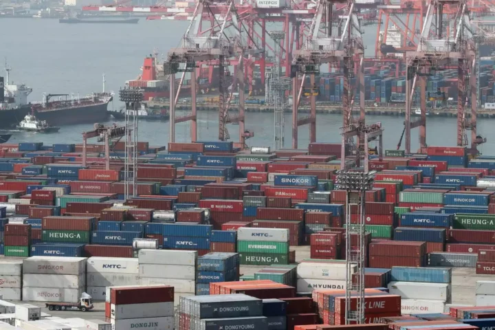 Containerhafen mit gestapelten Containern, Hafenkränen und Frachtschiffen im Hintergrund.