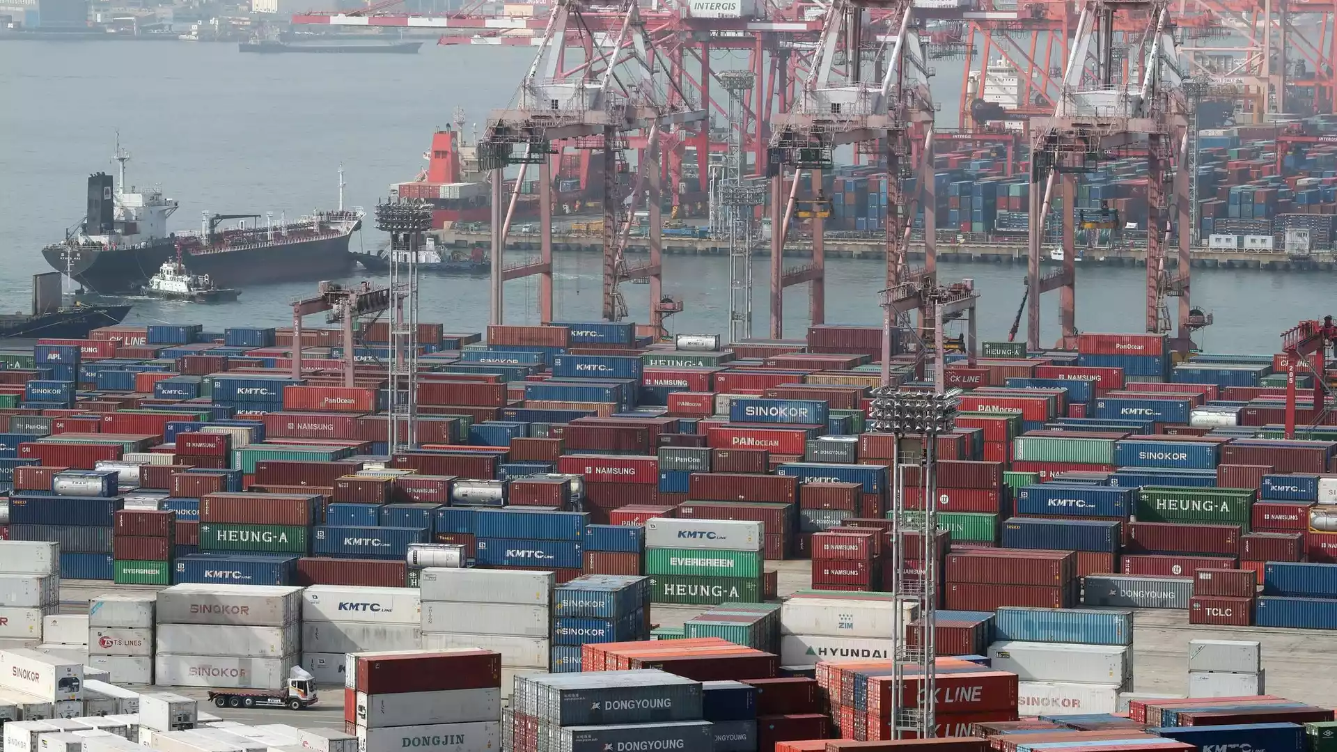 Containerhafen mit gestapelten Containern, Hafenkränen und Frachtschiffen im Hintergrund.