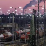 Güterzüge und Rangiergleise in einem beleuchteten Bahnbetriebswerk bei Abendhimmel.