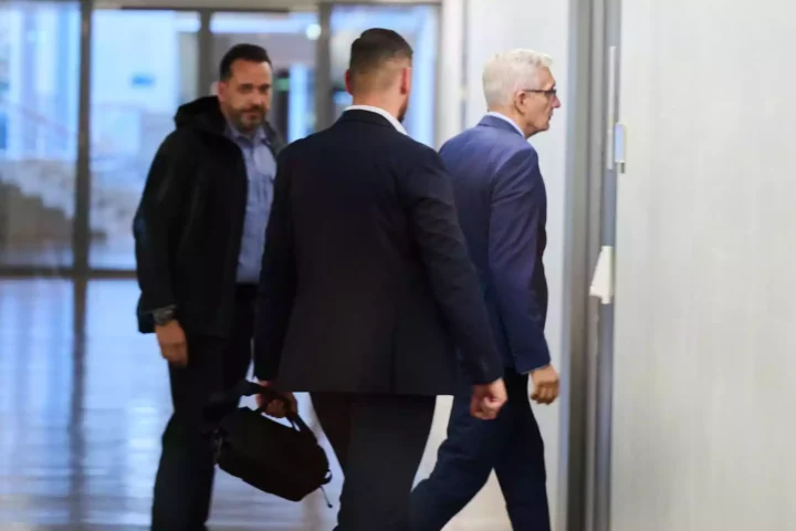 Drei Männer gehen durch einen hellen Flur, darunter ein älterer Mann im blauen Anzug mit weißen Haaren.