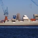 Eine Fregatte im Bau in einer Werft mit Kränen und Gerüst am Ufer.