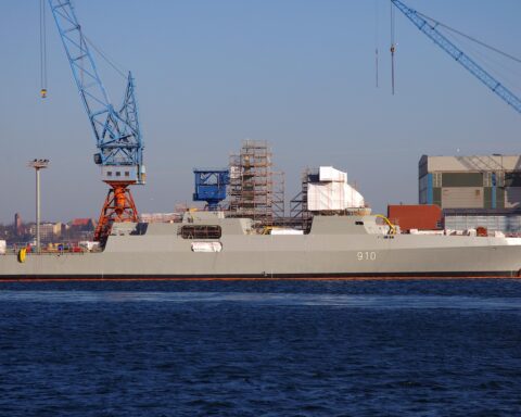Eine Fregatte im Bau in einer Werft mit Kränen und Gerüst am Ufer.