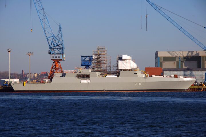 Eine Fregatte im Bau in einer Werft mit Kränen und Gerüst am Ufer.