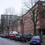 Mehrstöckiges rotes Bürogebäude mit parkenden Autos und kahlen Bäumen an einer Straße.