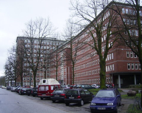Mehrstöckiges rotes Bürogebäude mit parkenden Autos und kahlen Bäumen an einer Straße.