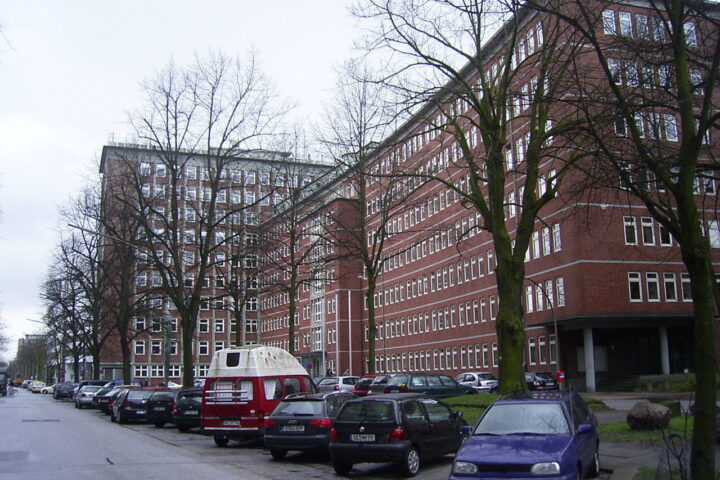 Mehrstöckiges rotes Bürogebäude mit parkenden Autos und kahlen Bäumen an einer Straße.