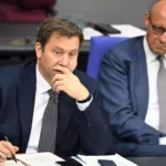 Zwei Politiker sitzen in einer Sitzung am Tisch, einer schreibt Notizen und blickt nach vorn.