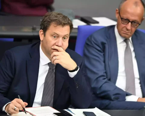 Zwei Politiker sitzen in einer Sitzung am Tisch, einer schreibt Notizen und blickt nach vorn.