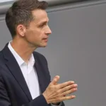 Andreas Audretsch im dunklen Anzug bei einer politischen Veranstaltung, im Profil und mit gefalteten Händen.