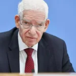 Josef Schuster sitzt im Anzug vor blauem Hintergrund und blickt ernst in die Kamera.