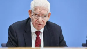 Josef Schuster sitzt im Anzug vor blauem Hintergrund und blickt ernst in die Kamera.