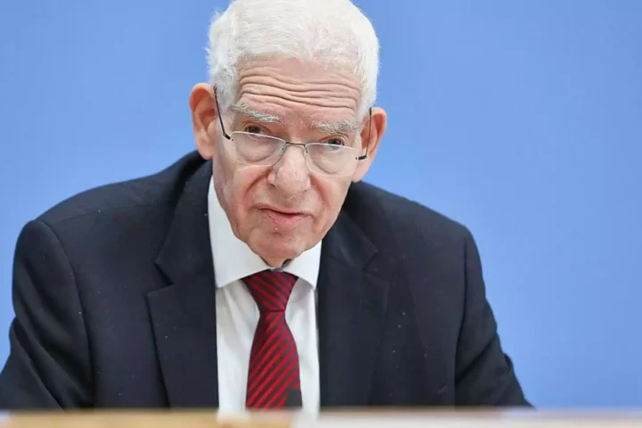 Josef Schuster sitzt im Anzug vor blauem Hintergrund und blickt ernst in die Kamera.