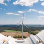 Blick von einer Windkraftanlage über Felder und Waldlandschaft unter blauem Himmel mit Wolken.