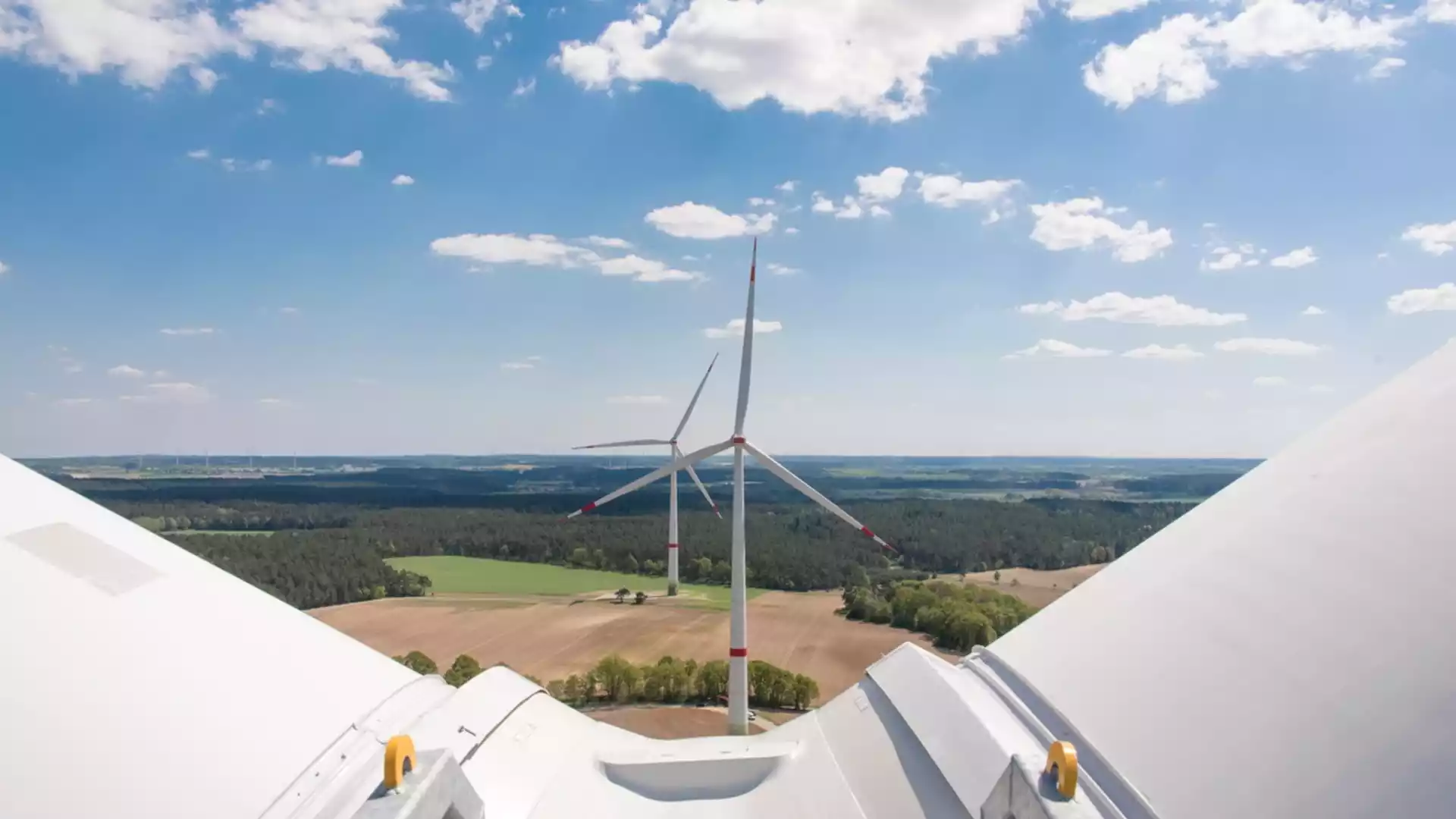 Blick von einer Windkraftanlage über Felder und Waldlandschaft unter blauem Himmel mit Wolken.
