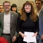 Mehrere Politikerinnen und Politiker stehen in einem Innenraum zusammen, eine Frau hält Papiere in der Hand.
