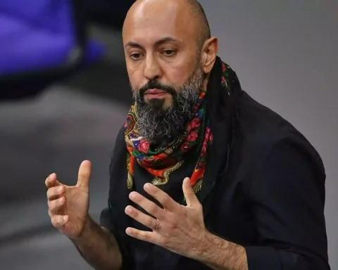 Ferat Koçak spricht mit erhobenen Händen vor grauem Hintergrund.