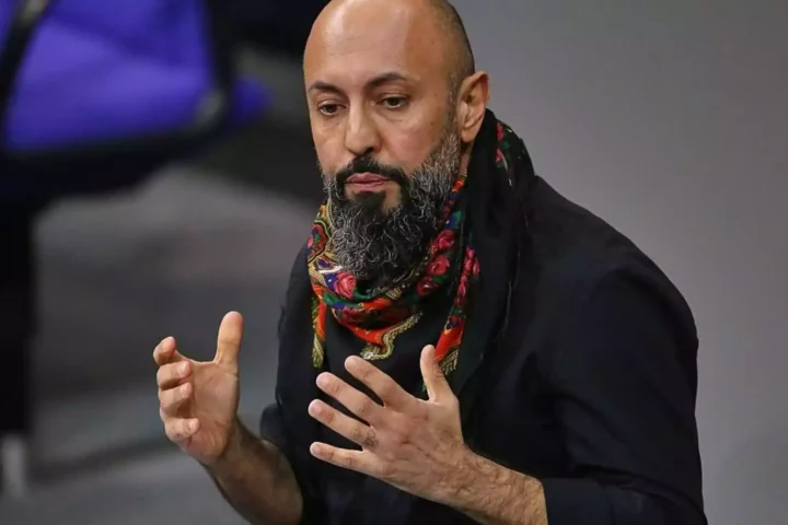 Ferat Koçak spricht mit erhobenen Händen vor grauem Hintergrund.