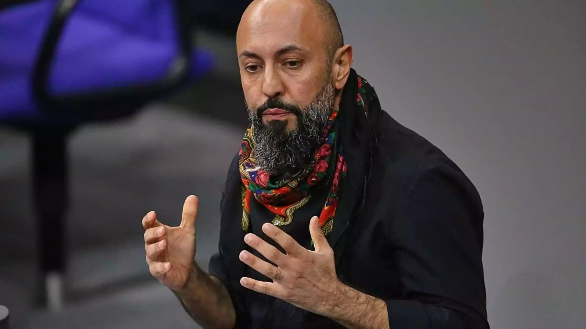 Ferat Koçak spricht mit erhobenen Händen vor grauem Hintergrund.