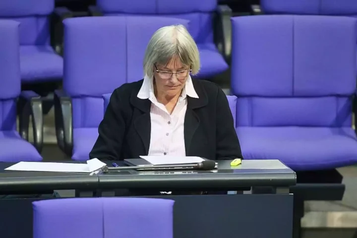Eine Frau sitzt in einem parlamentarisch wirkenden Saal vor violetten Sitzen und blickt auf Unterlagen vor sich.