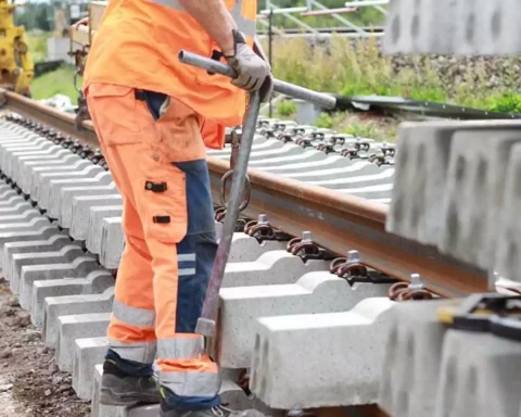 Ein Arbeiter in orangefarbener Warnkleidung arbeitet an einem Gleis mit Betonschwellen und Schienen.