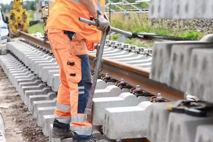 Ein Arbeiter in orangefarbener Warnkleidung arbeitet an einem Gleis mit Betonschwellen und Schienen.