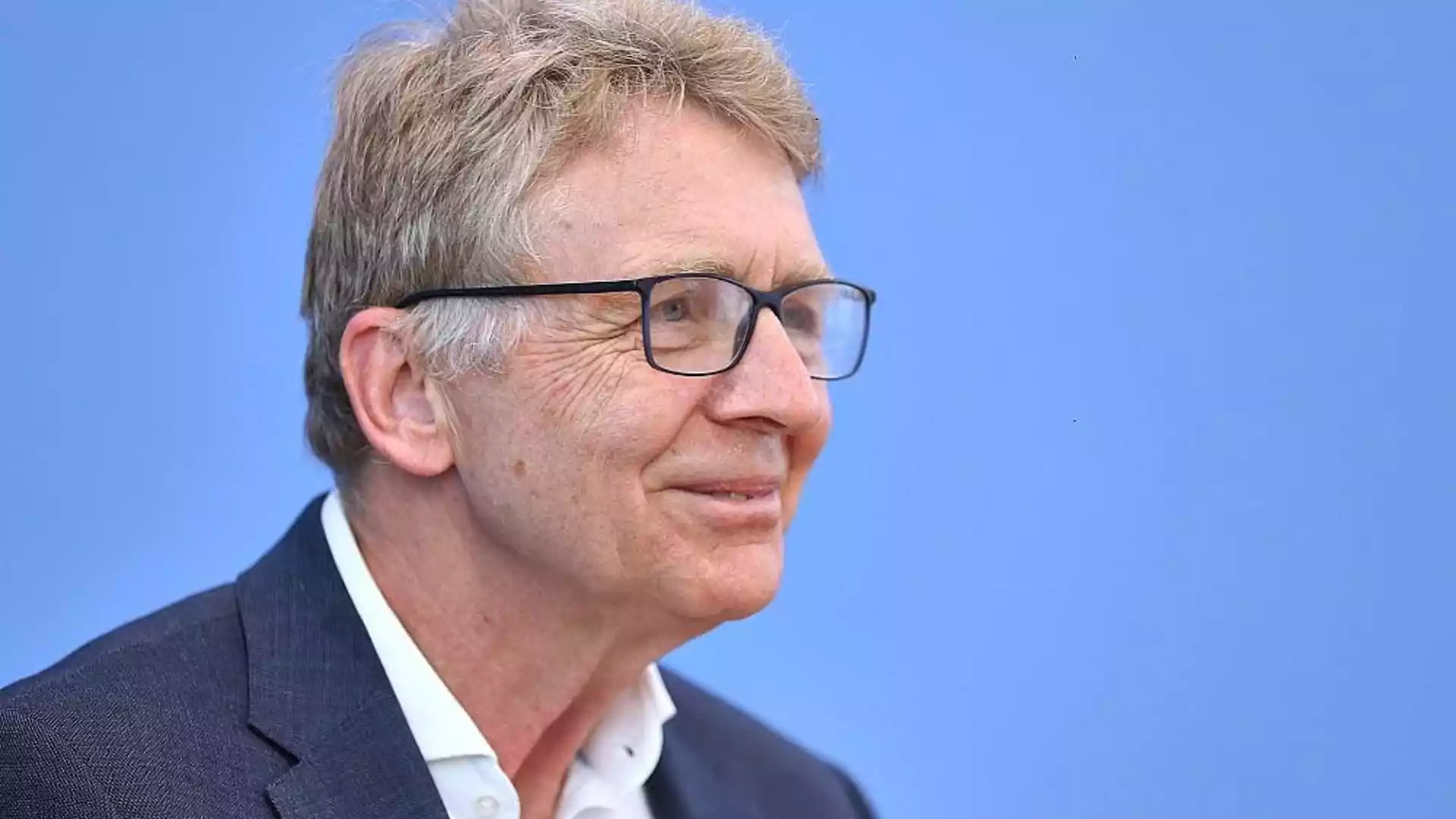 Porträt eines älteren Mannes mit Brille vor blauem Hintergrund.