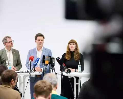 Drei Politiker stehen bei einer Pressekonferenz hinter mehreren Mikrofonen vor Publikum.