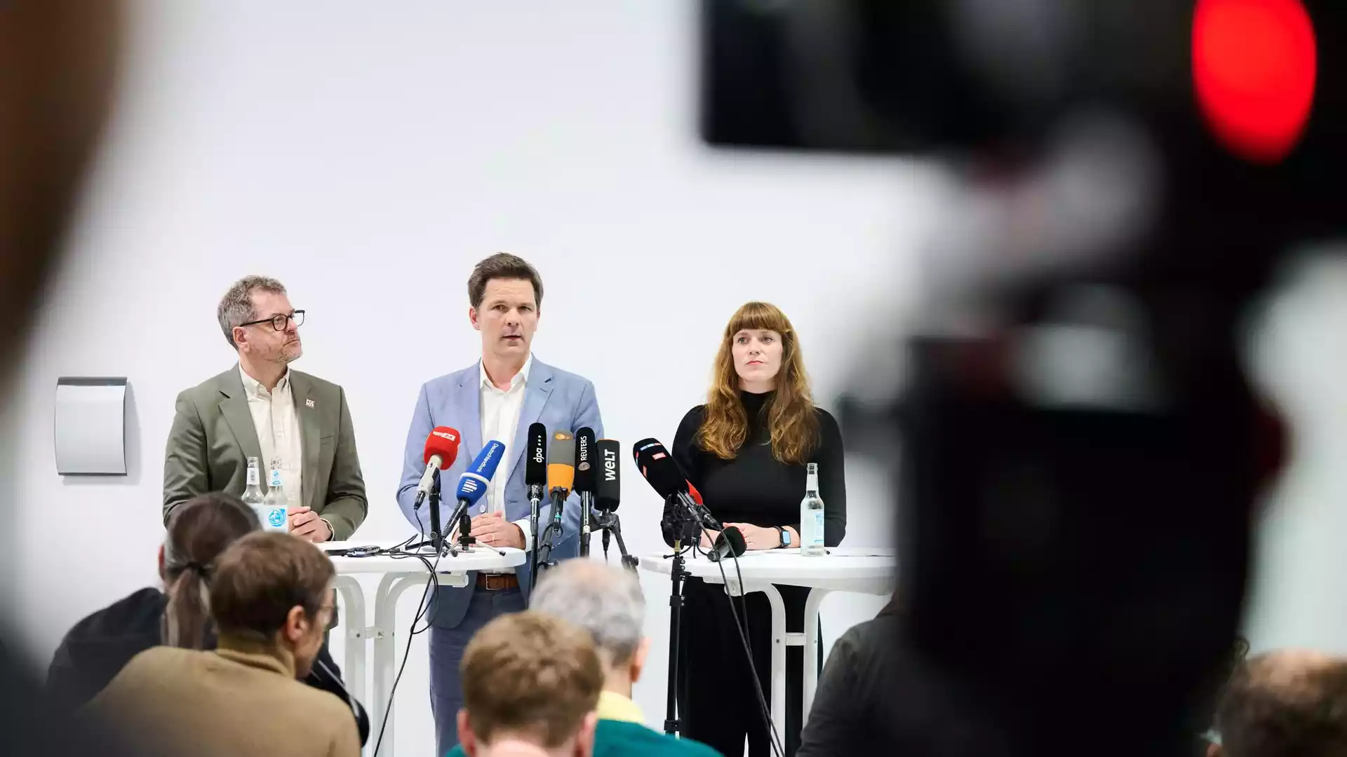 Drei Politiker stehen bei einer Pressekonferenz hinter mehreren Mikrofonen vor Publikum.