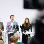 Drei Personen sprechen bei einer Pressekonferenz vor mehreren Mikrofonen in einem hellen Raum.