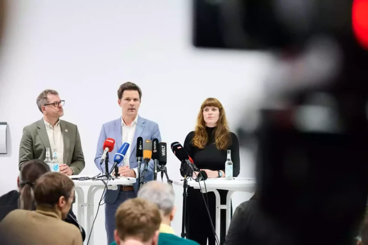 Drei Personen sprechen bei einer Pressekonferenz vor mehreren Mikrofonen in einem hellen Raum.