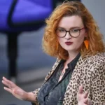 Eine Frau mit rotem, lockigem Haar und Brille spricht gestikulierend in einem Saal.