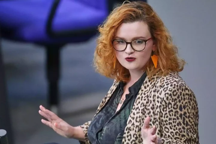 Eine Frau mit rotem, lockigem Haar und Brille spricht gestikulierend in einem Saal.