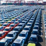 Reihen neuer Autos auf einem großen Hafengelände mit Containern und Kränen im Hintergrund.