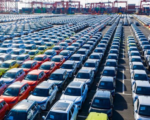 Reihen neuer Autos auf einem großen Hafengelände mit Containern und Kränen im Hintergrund.