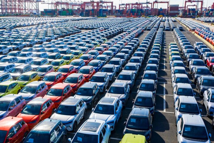 Reihen neuer Autos auf einem großen Hafengelände mit Containern und Kränen im Hintergrund.